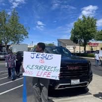 Church member holding a Jesus is Resurrection and Life sign near parked cars